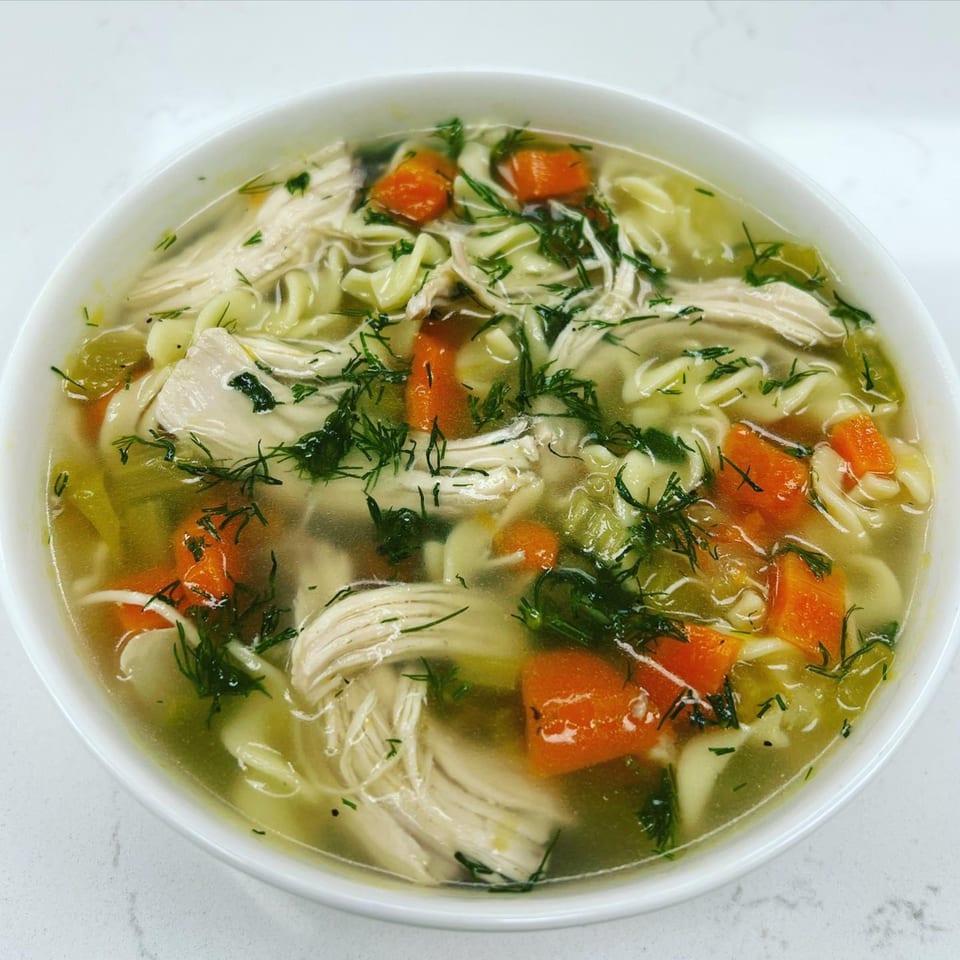 A white bowl of Mike’s Gourmet Deli chicken noodle soup with carrots, celery, and dill.