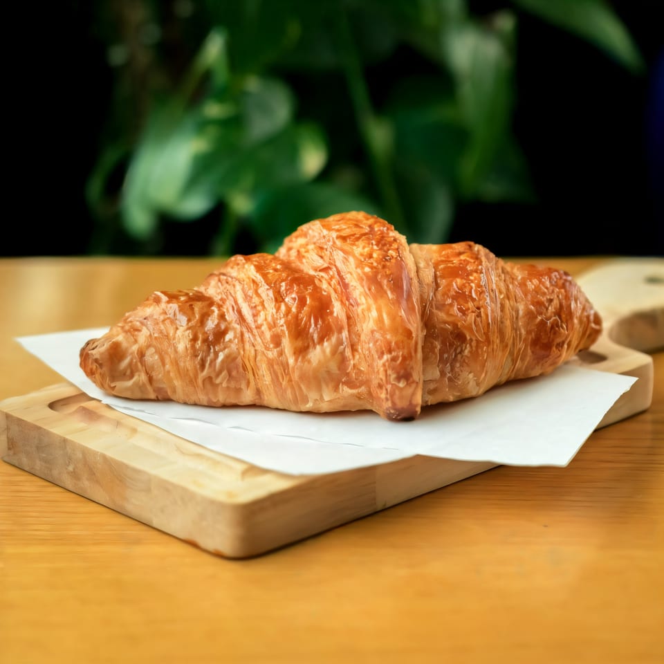 A golden-brown croissant rests on white paper and a wooden board, with green foliage in the background.